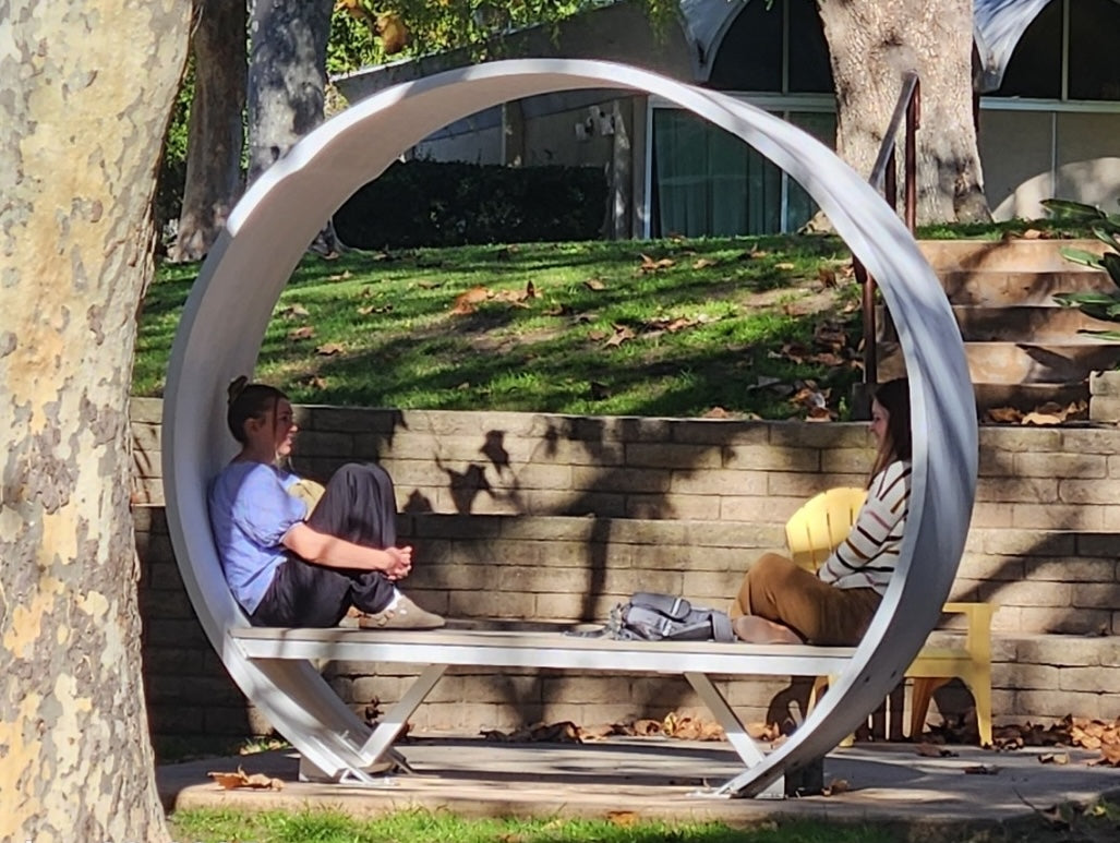 Two students talking on a Noblewins shaded bench made from a repurposed wind turbine blade in a campus setting