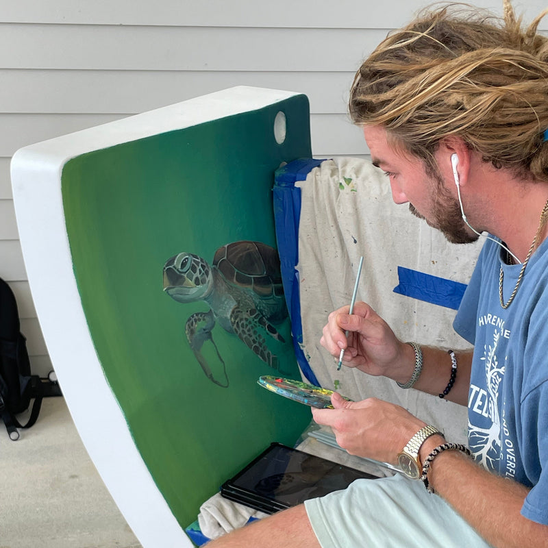 Artist hand-painting a detailed sea turtle mural on a primed Noblewins bench made from a repurposed wind turbine blade