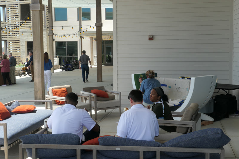 Artist painting a public art bench made from a repurposed wind turbine blade while people gather in an outdoor courtyard event space